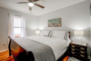 A bedroom with a bed, a ceiling fan, a window with curtains, a bedside lamp, a dresser, and a painting on the wall. - Enjoy Staunton in the Heart of Shenandoah Valley (Staunton)