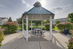 Ein Gazebo mit Metallveranda und weißen Säulen, umgeben von einer gepflasterten Fläche mit Picknicktisch und Bänken.