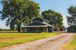 A two-story house with a porch, surrounded by a grassy field and large trees.