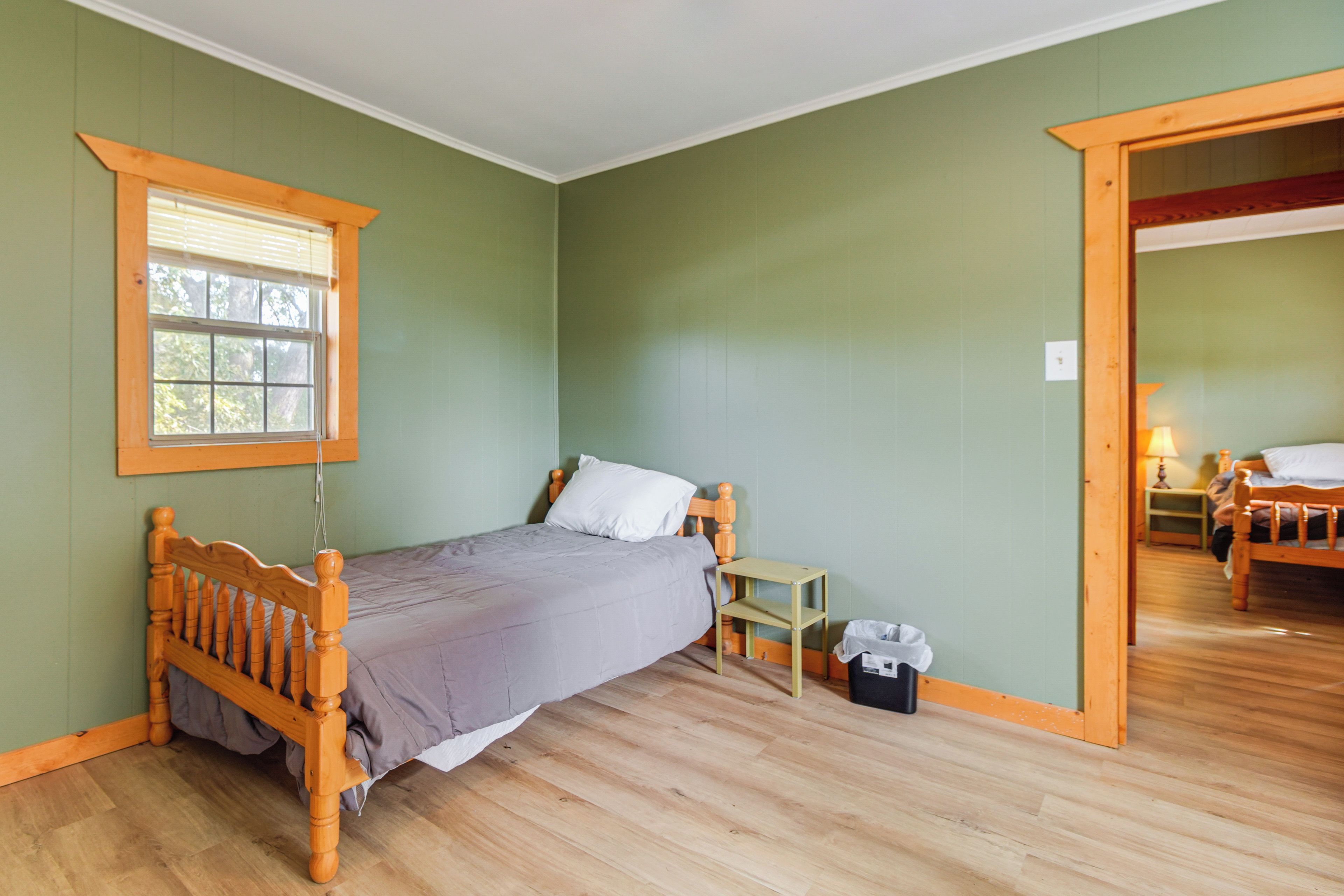 Un dormitorio con cama de madera, una mesita de noche pequeña, una ventana con persianas y una puerta que da a otra habitación.