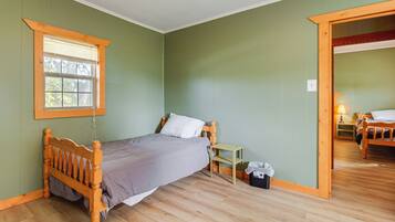 Un dormitorio con cama de madera, una mesita de noche pequeña, una ventana con persianas y una puerta que da a otra habitación.