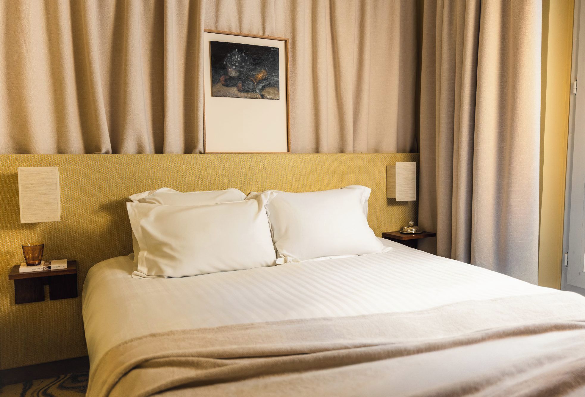 Una habitación de hotel con cama, dos almohadas, una mesita de noche con lámpara y un cuadro en la pared.