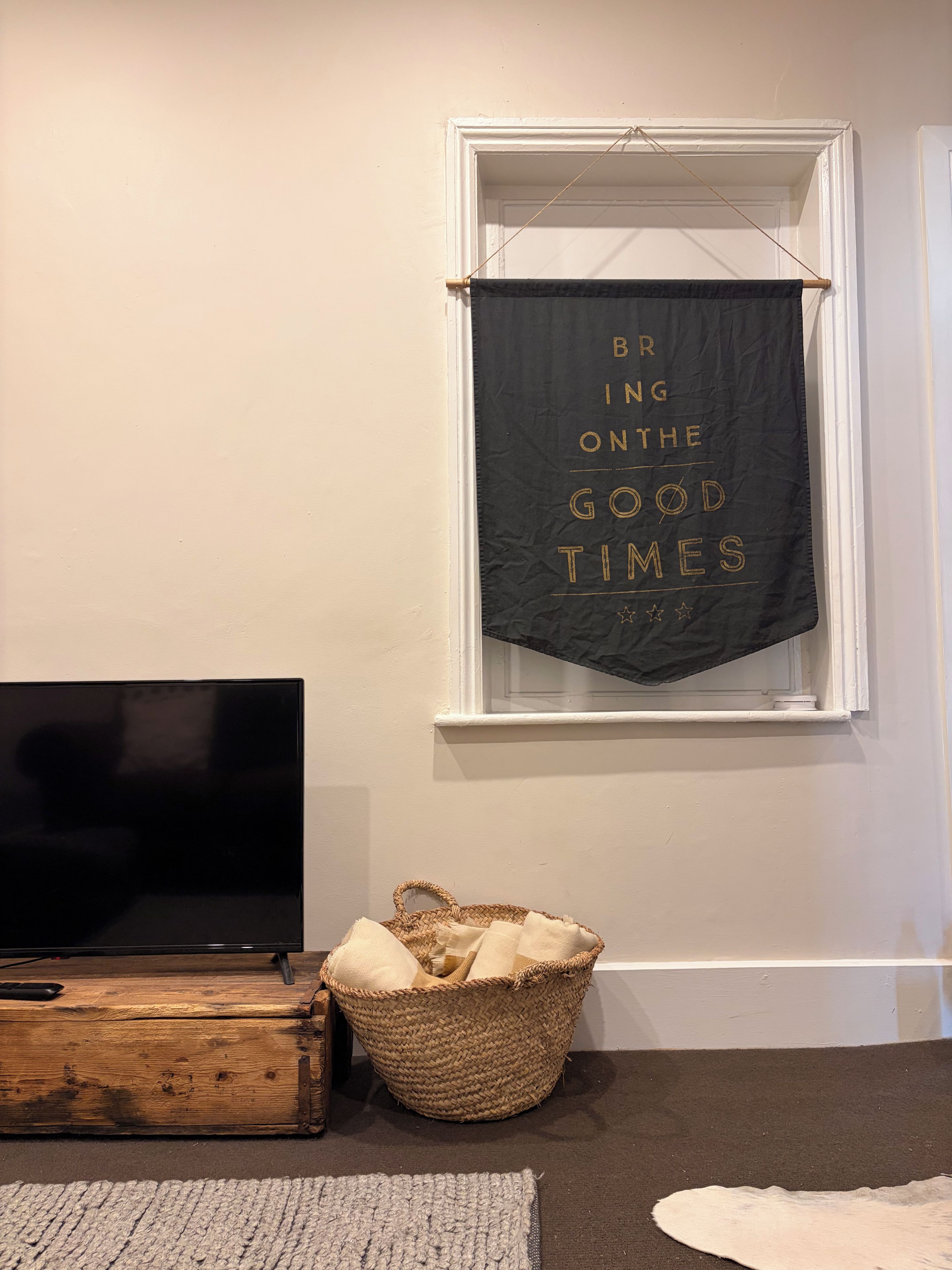 Una habitación con televisión, una cesta tejida, una alfombra y una ventana con un banderín colgado.