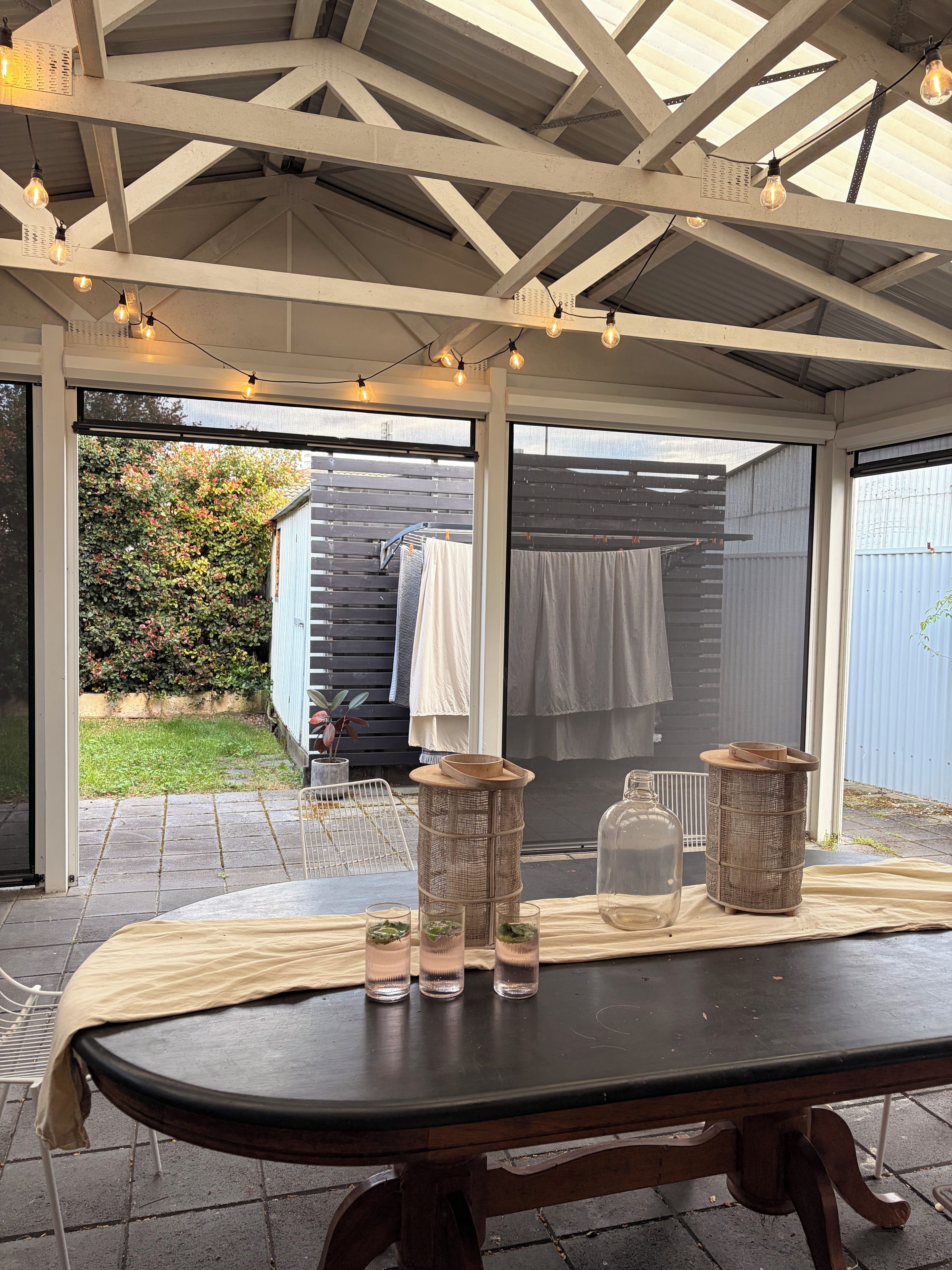 A covered outdoor area with a table set for dining, string lights overhead, and a view of a garden through the open sides.