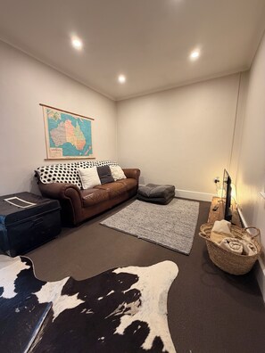 A living room with a brown leather sofa, a map of Australia on the wall, a black suitcase, a woven basket, and a remote control. - Rêverie | cosy coastal cottage, walking distance to cafes and beach (Kingston SE)