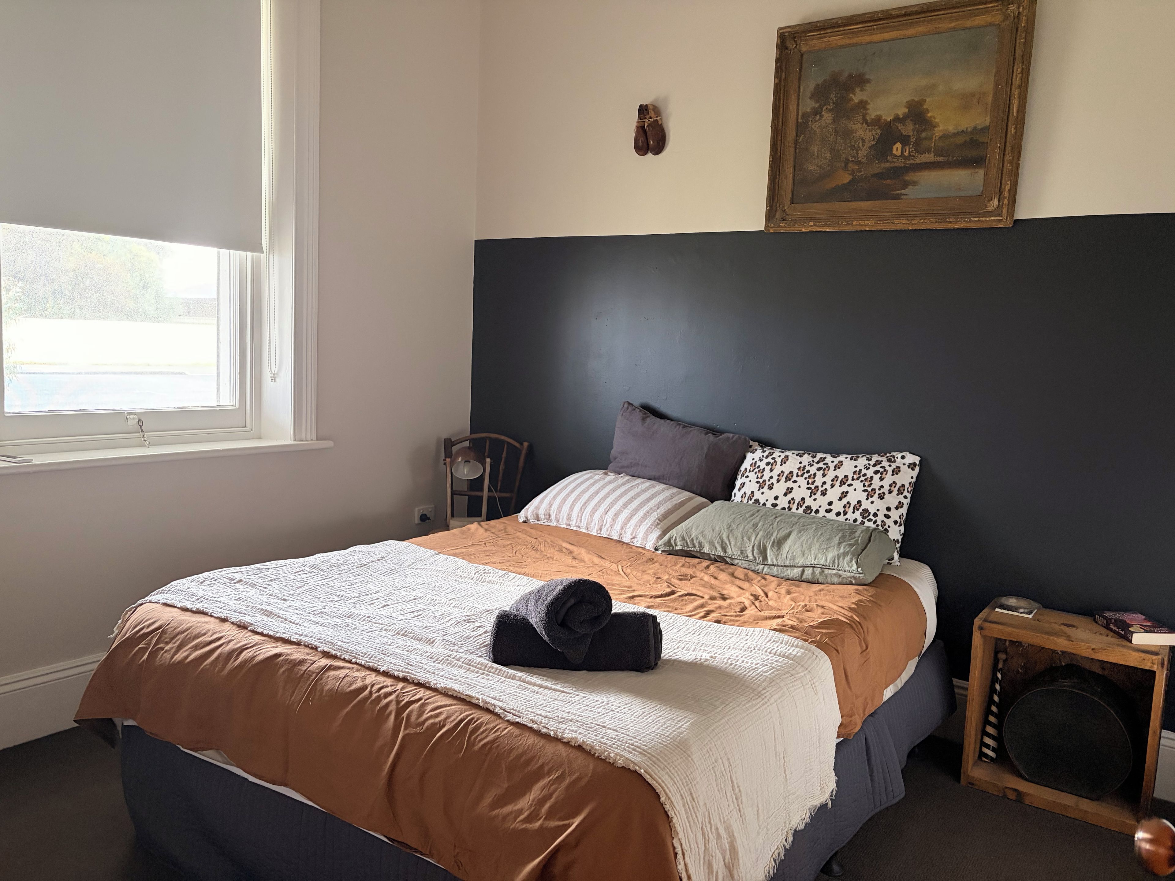 Un dormitorio con cama, ventana, una silla, una mesita con un libro y un cuadro en la pared.