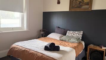 Un dormitorio con cama, ventana, una silla, una mesita con un libro y un cuadro en la pared.