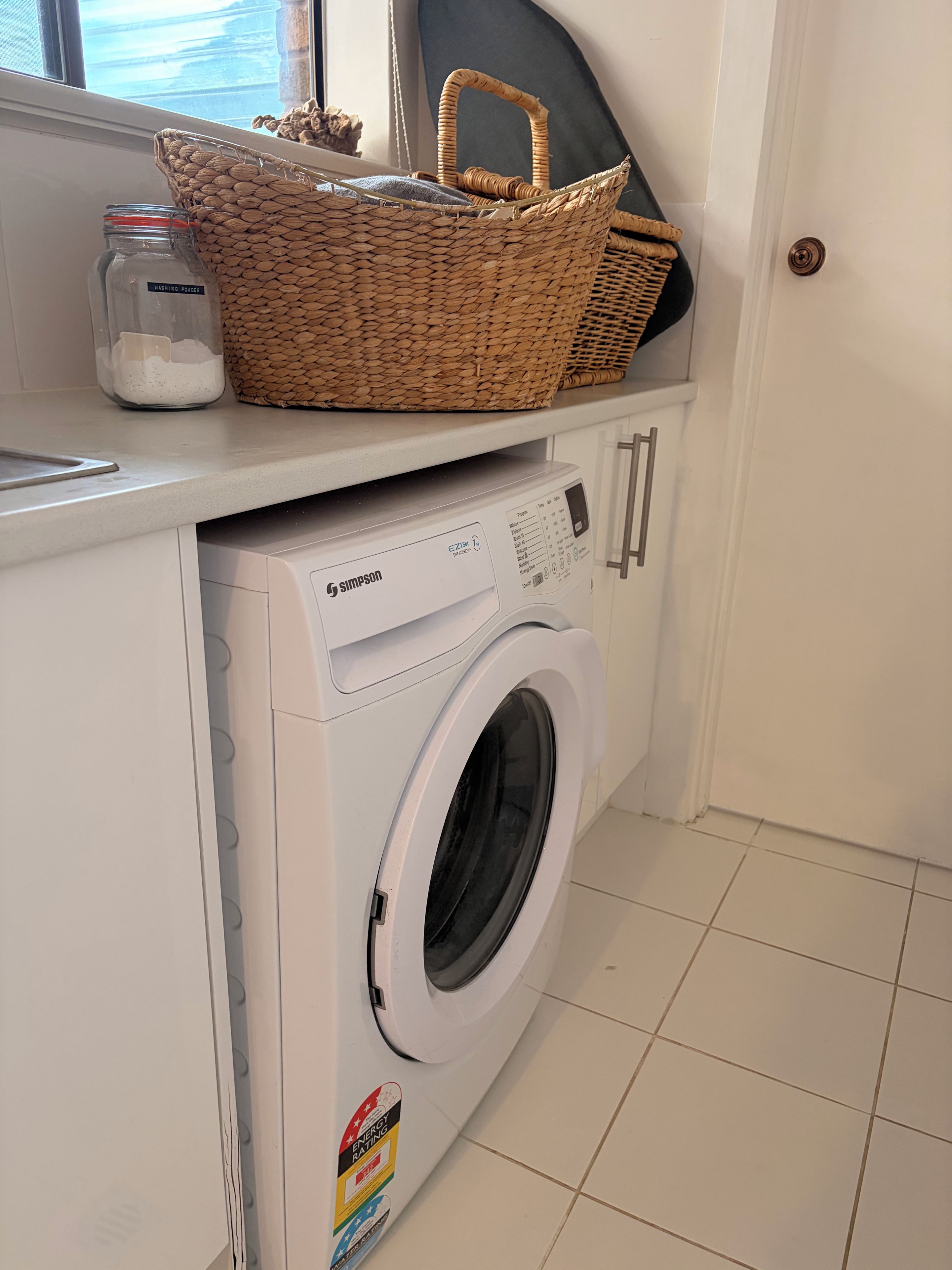 A white washing machine with a control panel, a wicker basket, and a jar labeled 'washing powder' on a countertop.