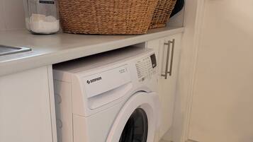 A white washing machine with a control panel, a wicker basket, and a jar labeled 'washing powder' on a countertop.
