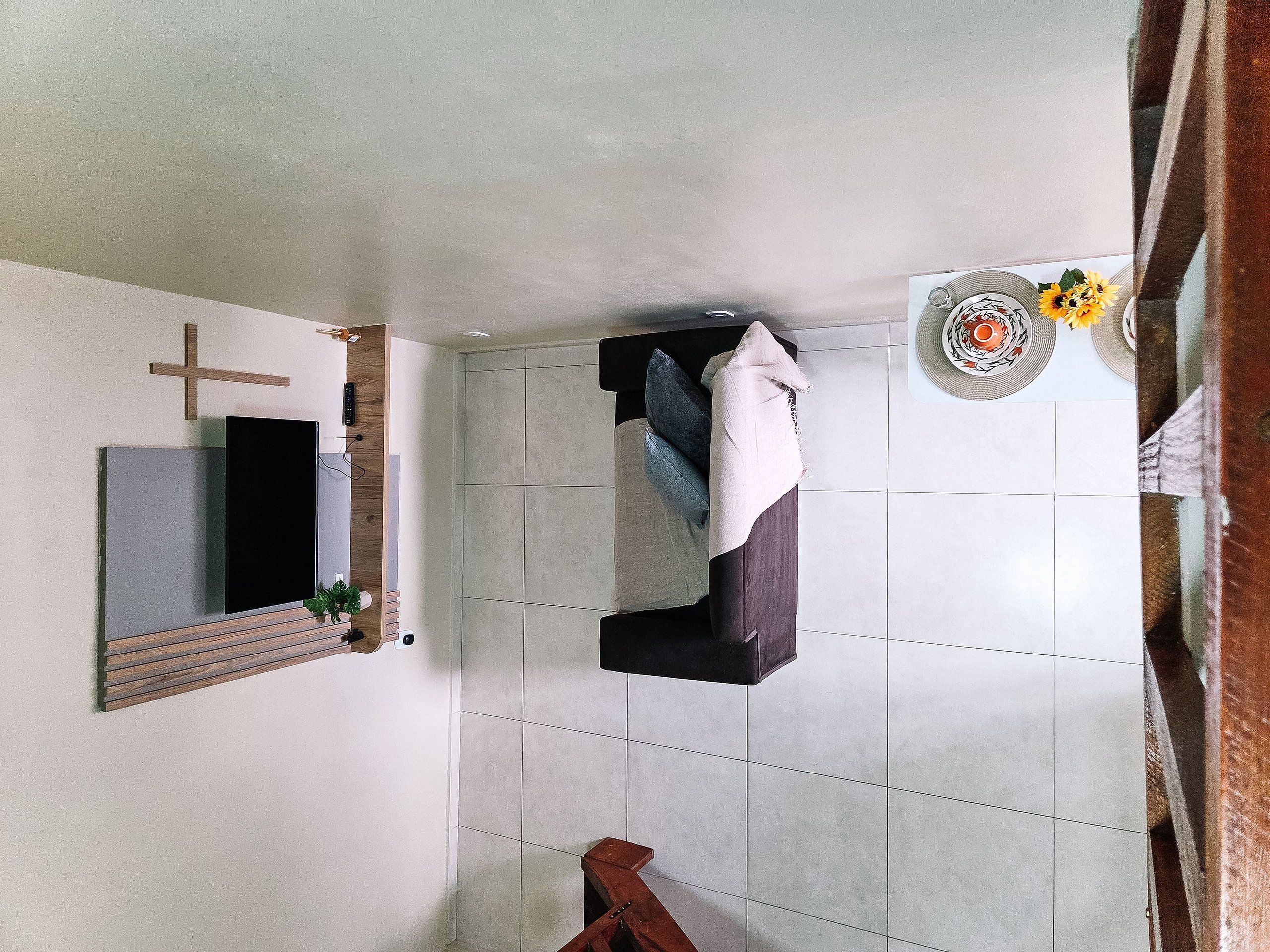 Quarto com cama, uma cruz na parede, uma planta pequena e uma mesa com um prato decorativo.