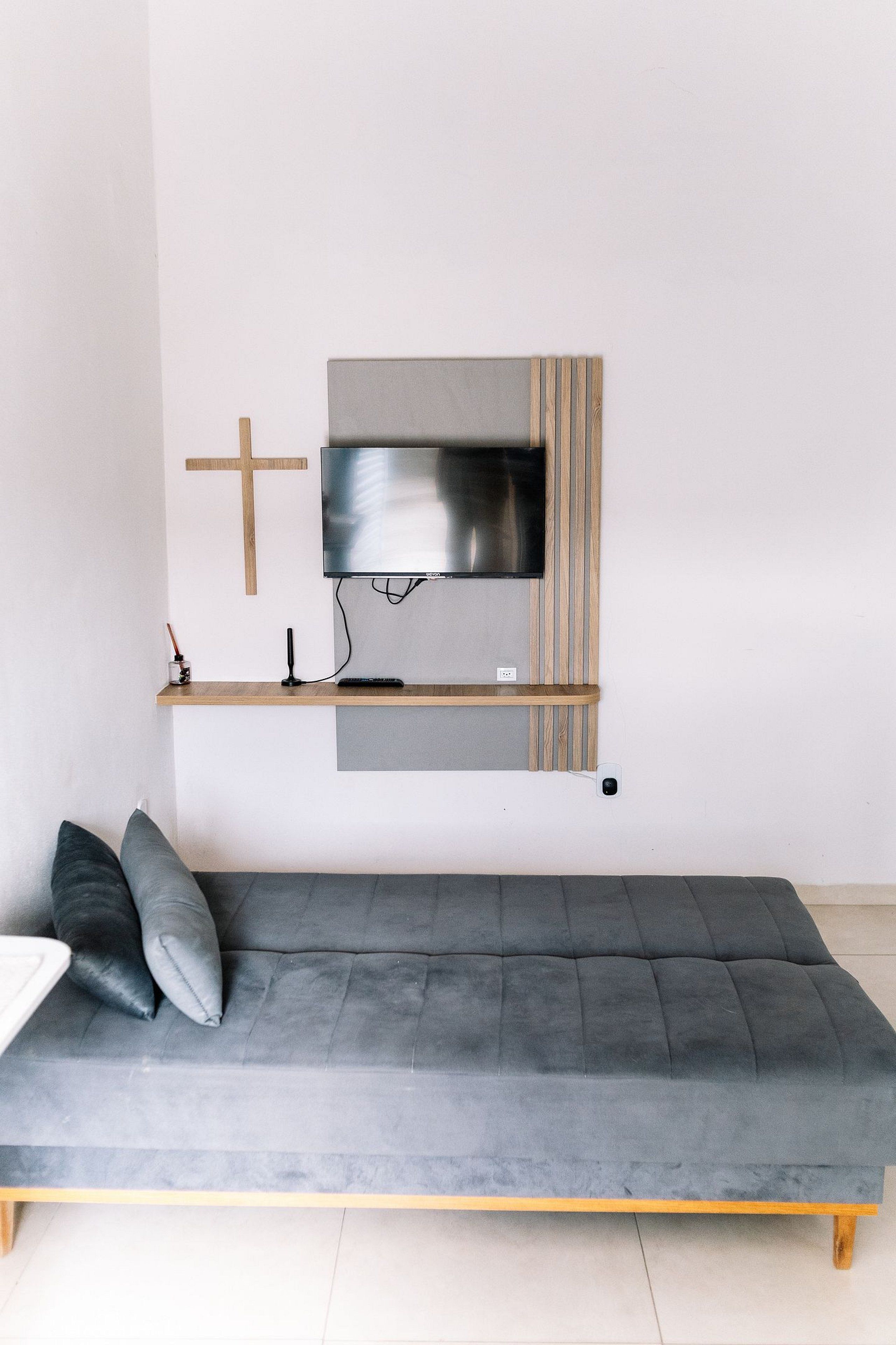 Une pièce au décor minimaliste, avec un canapé gris, un téléviseur fixé au mur et une étagère en bois ornée d’une croix.
