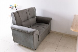 Un sillón gris con un respaldo alto y reposabrazos, ubicado en una habitación con piso de baldosas y pared blanca.