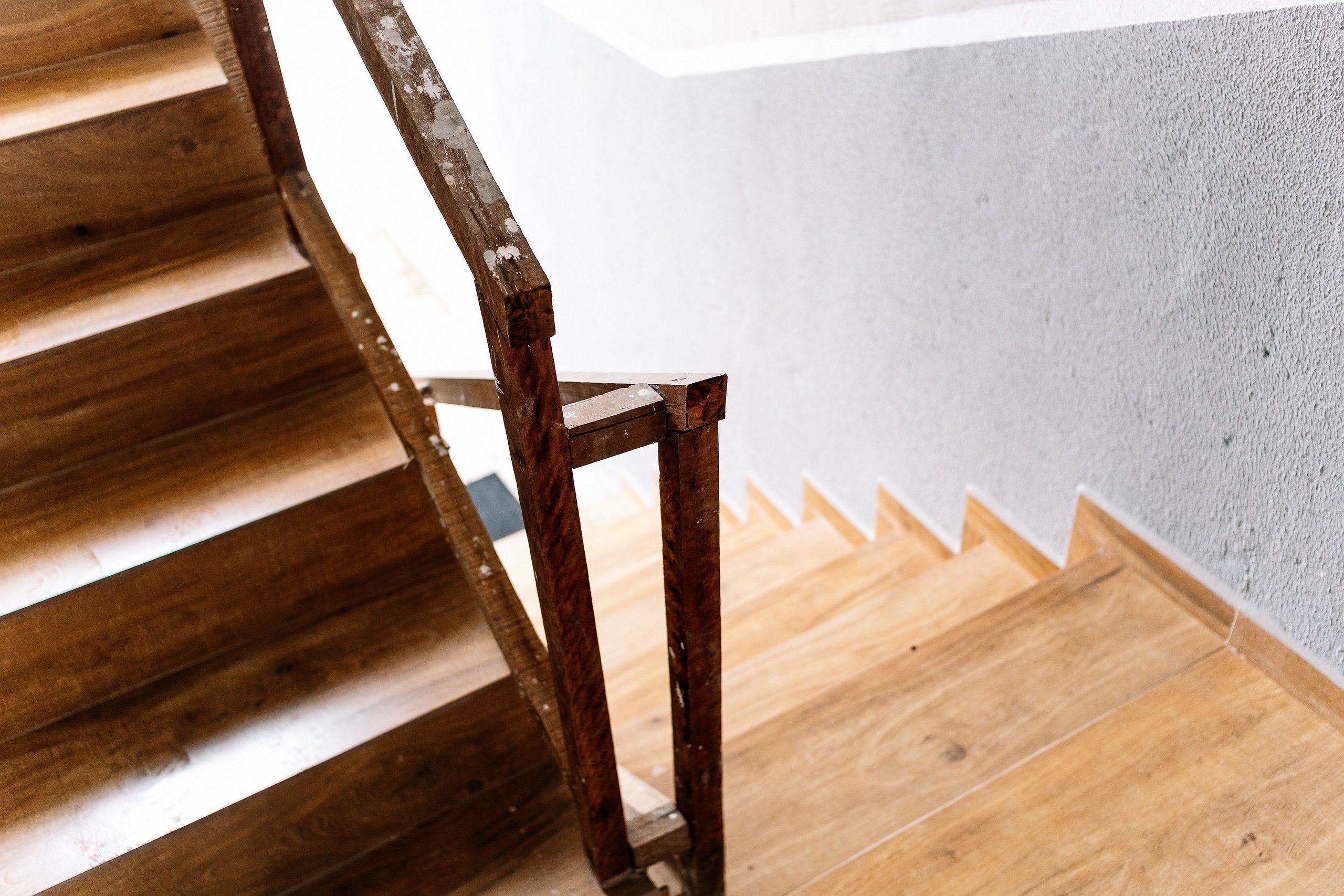 A wooden staircase with a metal handrail leading to a room with a white wall and a wooden floor.