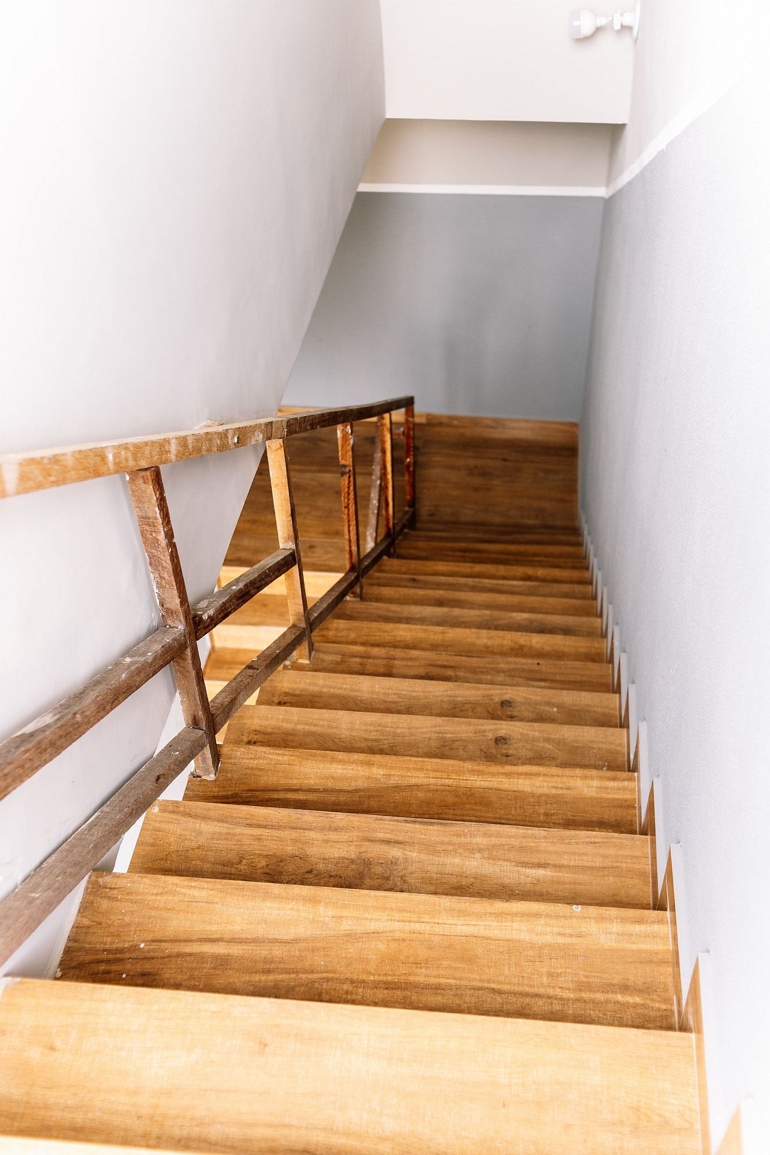 A wooden staircase with a metal handrail, leading to a landing with a white wall and a light switch.