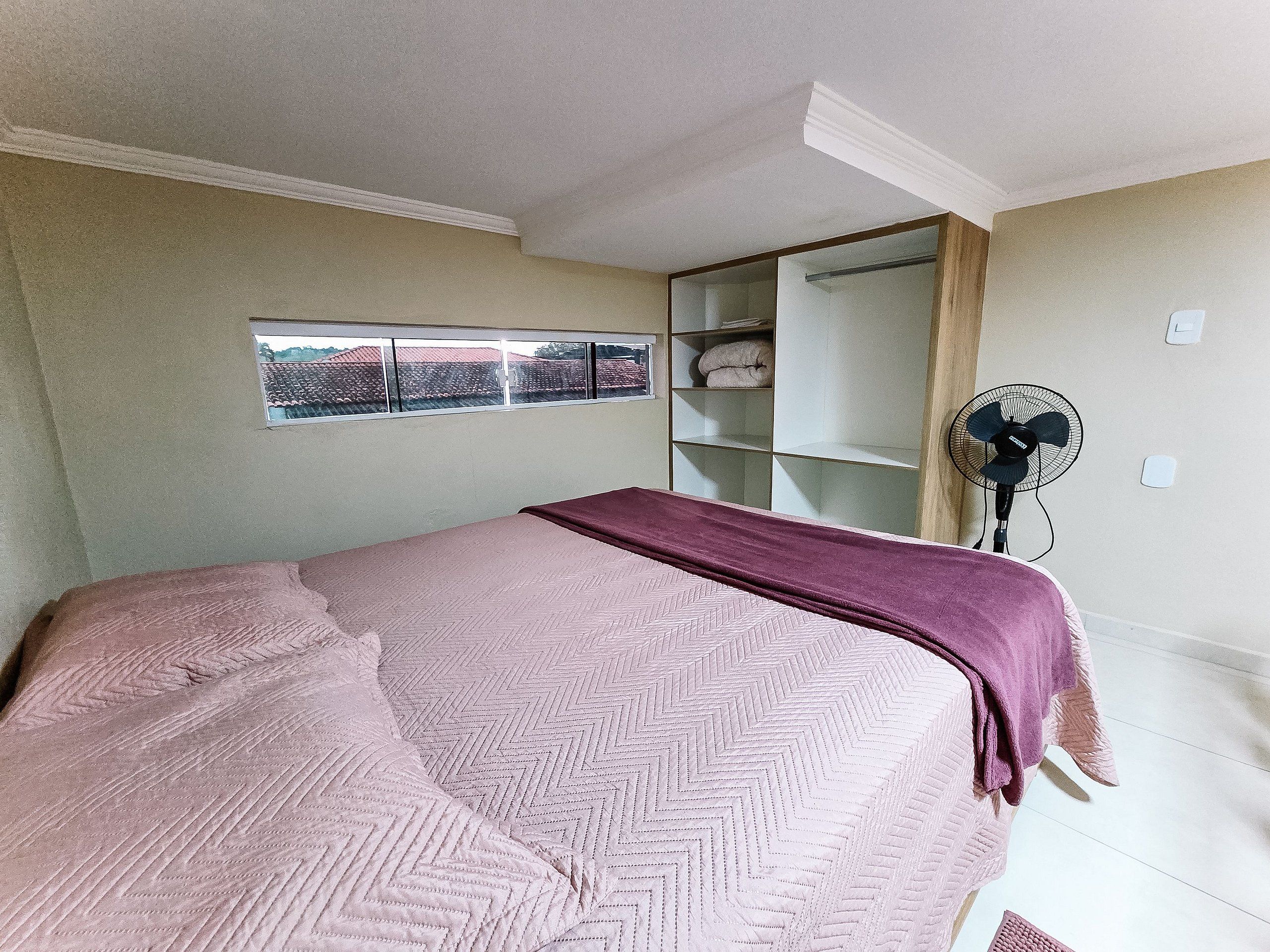 A bedroom with a bed, a window, a wardrobe, and a standing fan.