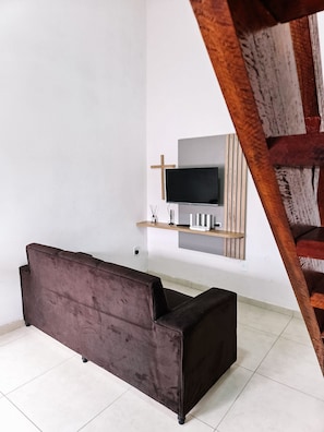 Una habitación con un sofá marrón oscuro, un televisor de pantalla plana montado en un estante de madera y una escalera al fondo.