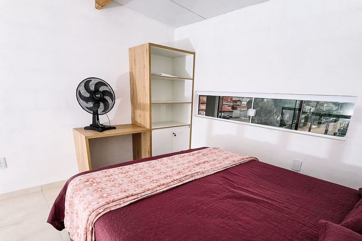 A bedroom with a bed, a wooden nightstand, a fan, a window with a view, and a red rug.