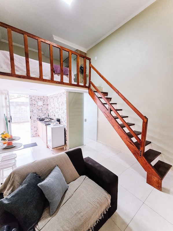 A living room with a sofa, a dining table with chairs, a kitchen area, and a wooden staircase.
