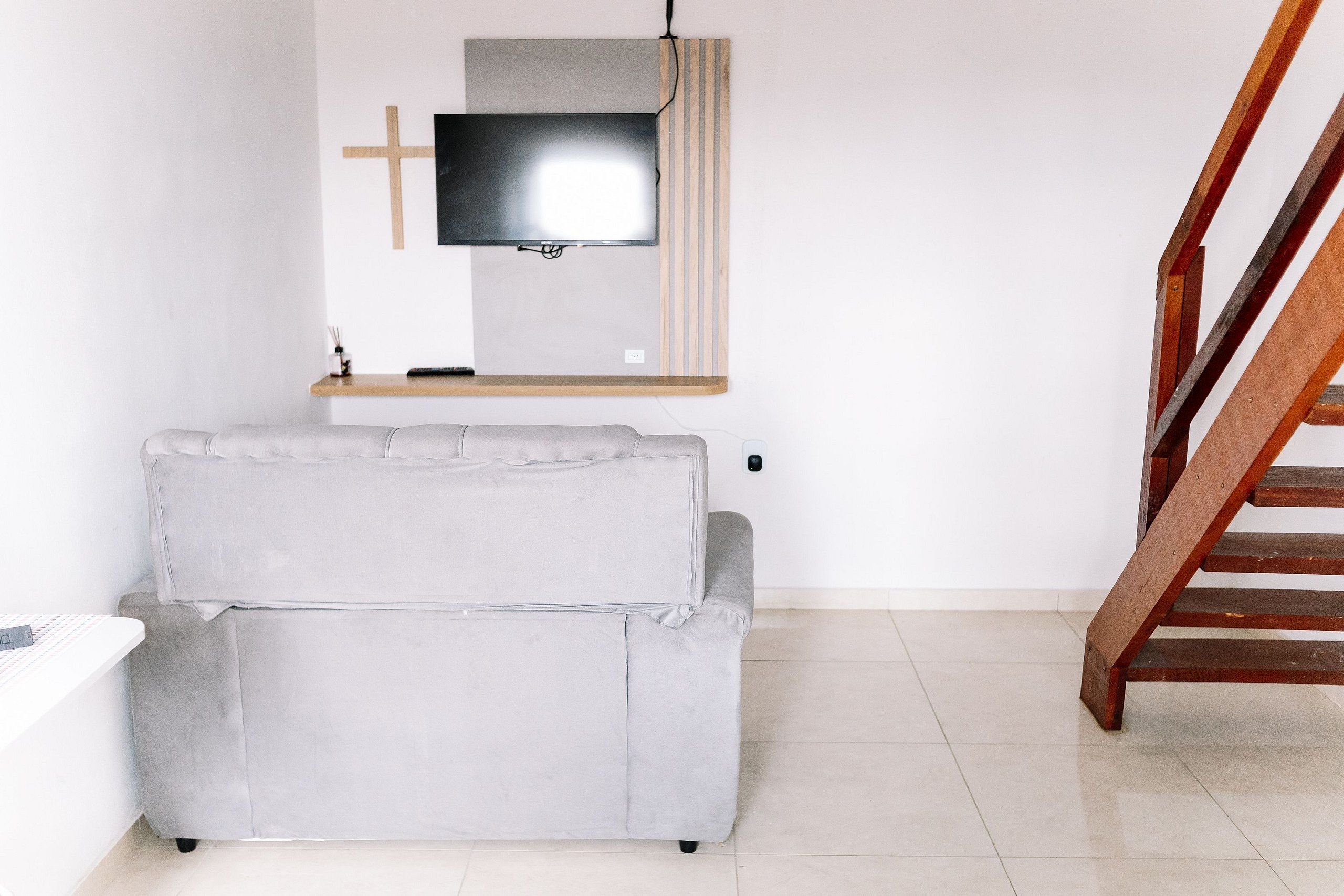 Une pièce avec un canapé gris, un téléviseur fixé au mur, une étagère en bois et un escalier en bois.
