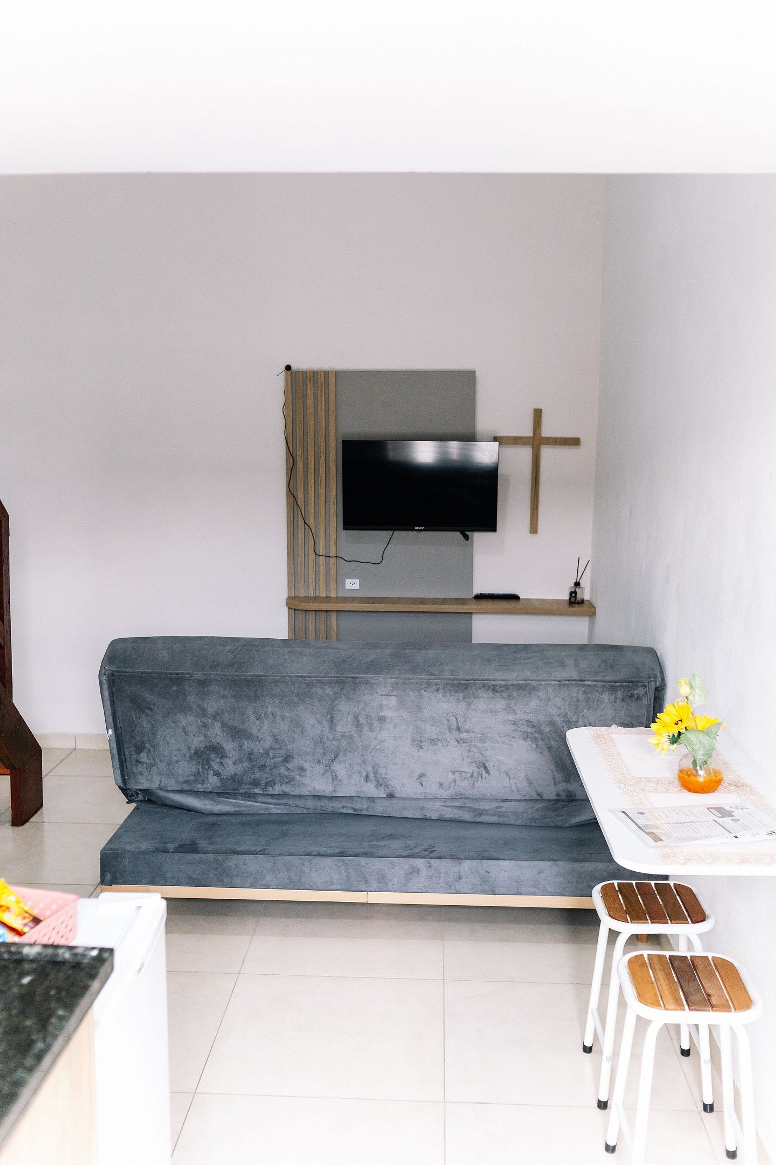 Una sala de estar moderna con un sofá gris, un televisor de pantalla plana montado en un panel de madera, una pequeña mesa blanca con un jarrón de flores y un par de taburetes blancos.