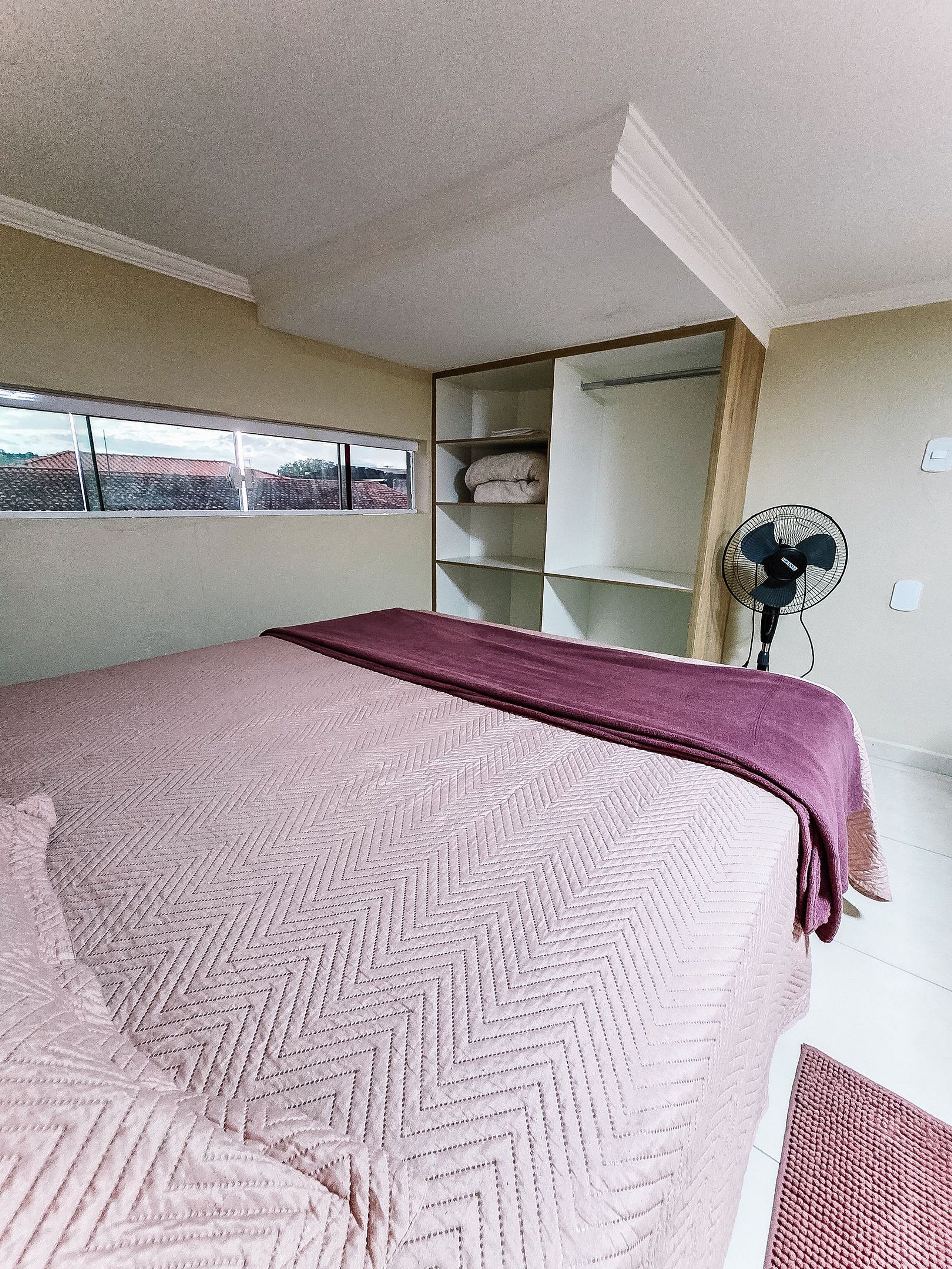 Ein Schlafzimmer mit einem Bett, einem Fenster, einem Ventilator und einem Regal.
