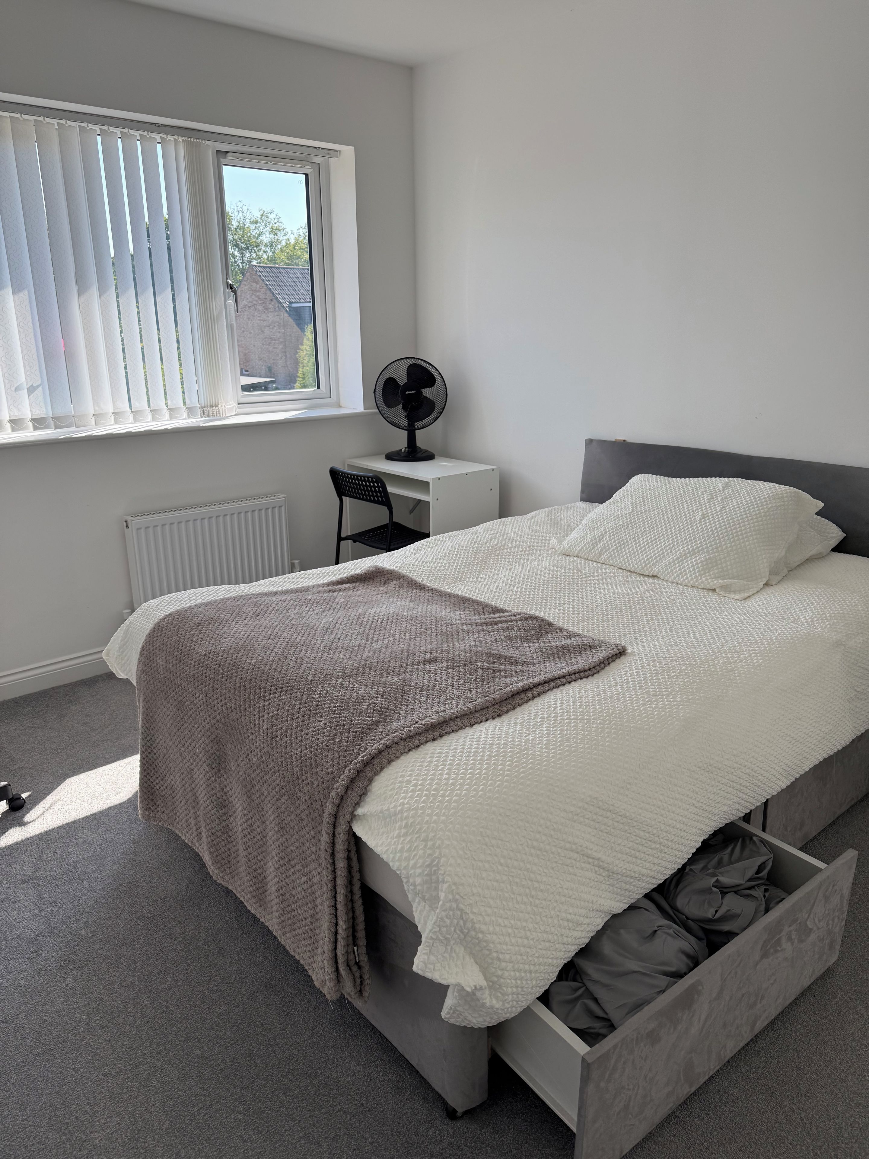 A bedroom with a bed, a desk, a chair, a radiator, and a window with blinds.