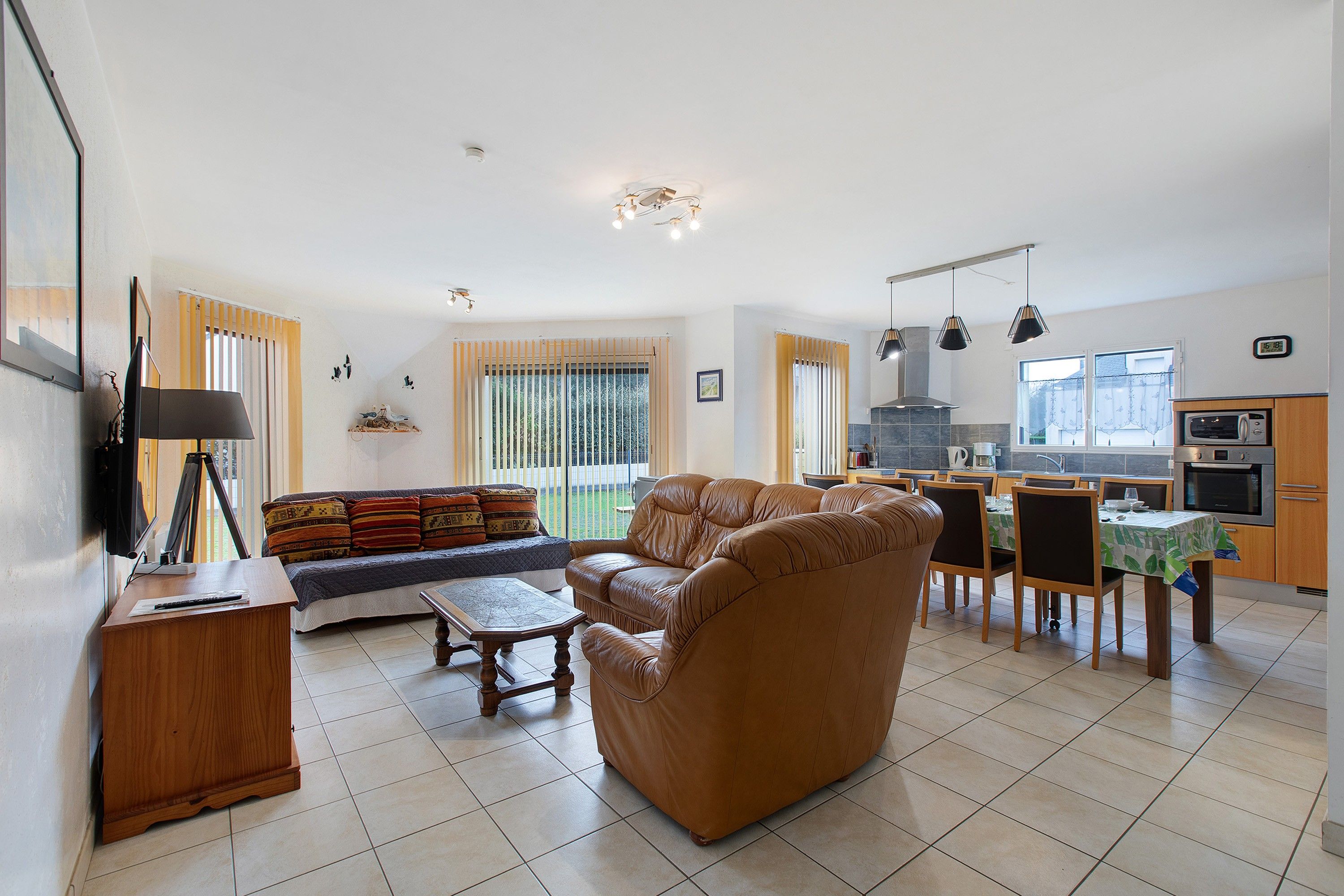 En moderne stue med sofa, spisebord og stole, et køkken med hårde hvidevarer, og et stort vindue med gardiner.