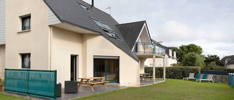 Une maison à deux étages avec un toit en pente, une terrasse couverte et un jardin clôturé.