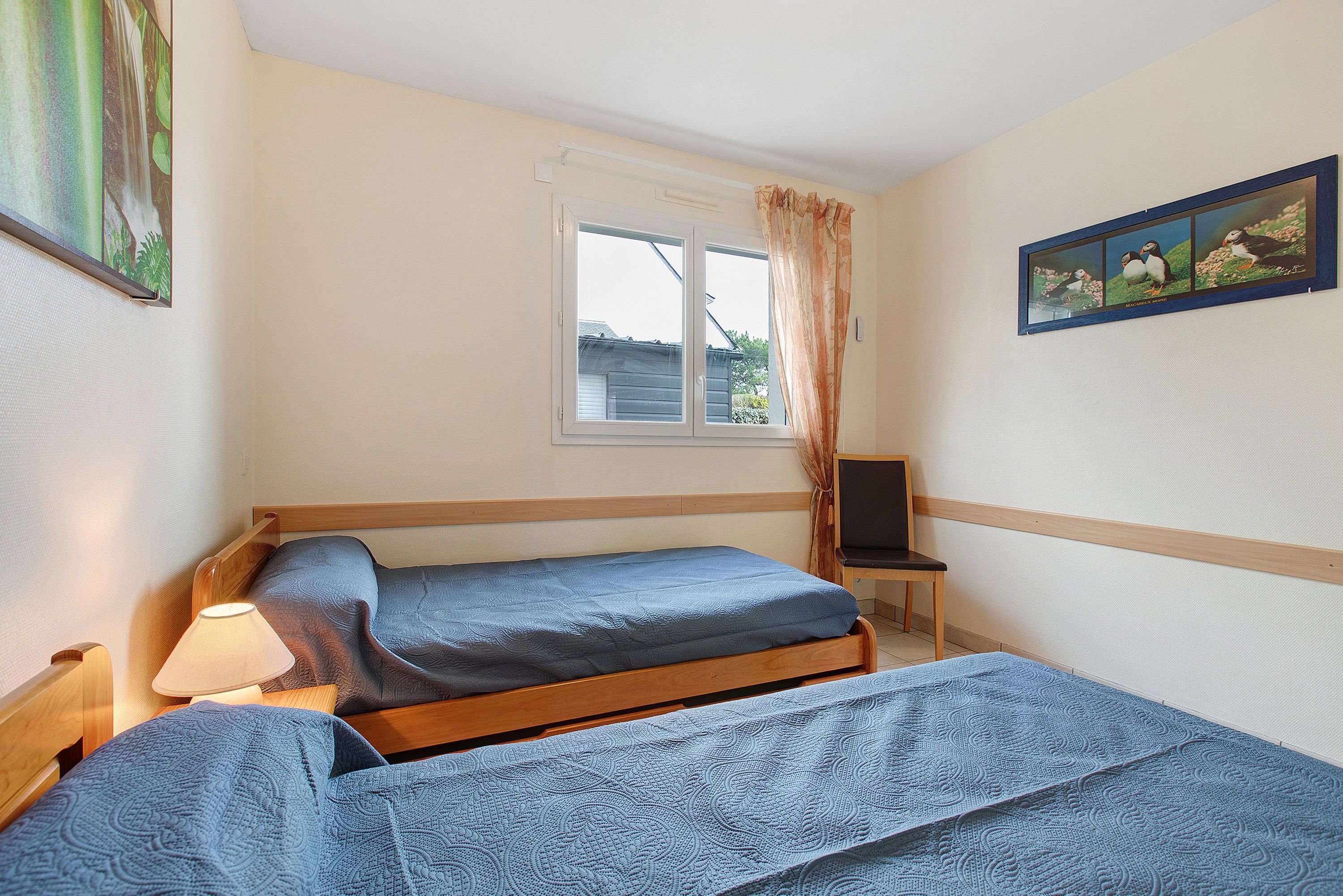 A room with two beds, a window with curtains, a small table with a lamp, and a framed picture on the wall.