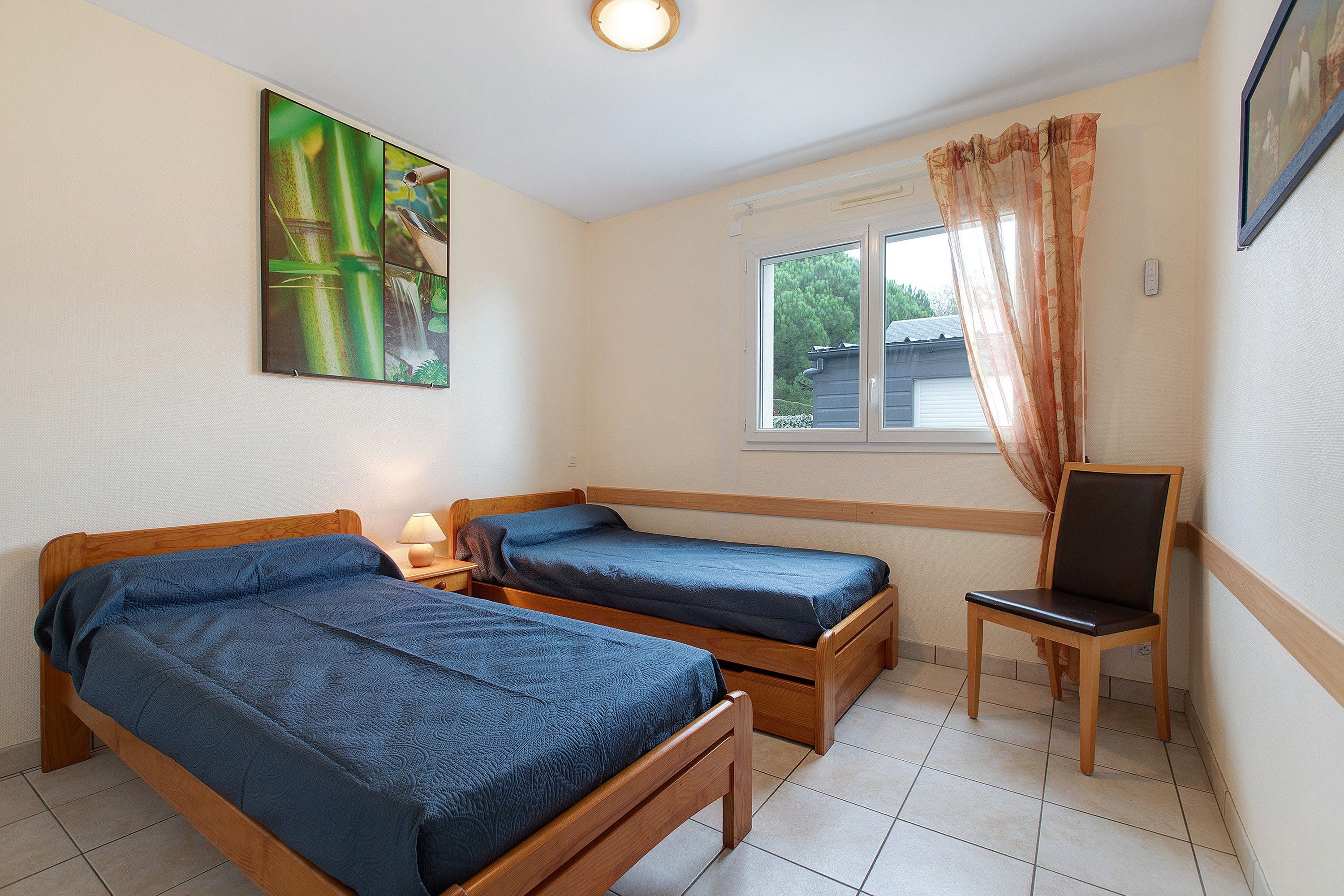 A room with two wooden beds, a chair, a window with curtains, and a wall-mounted picture.