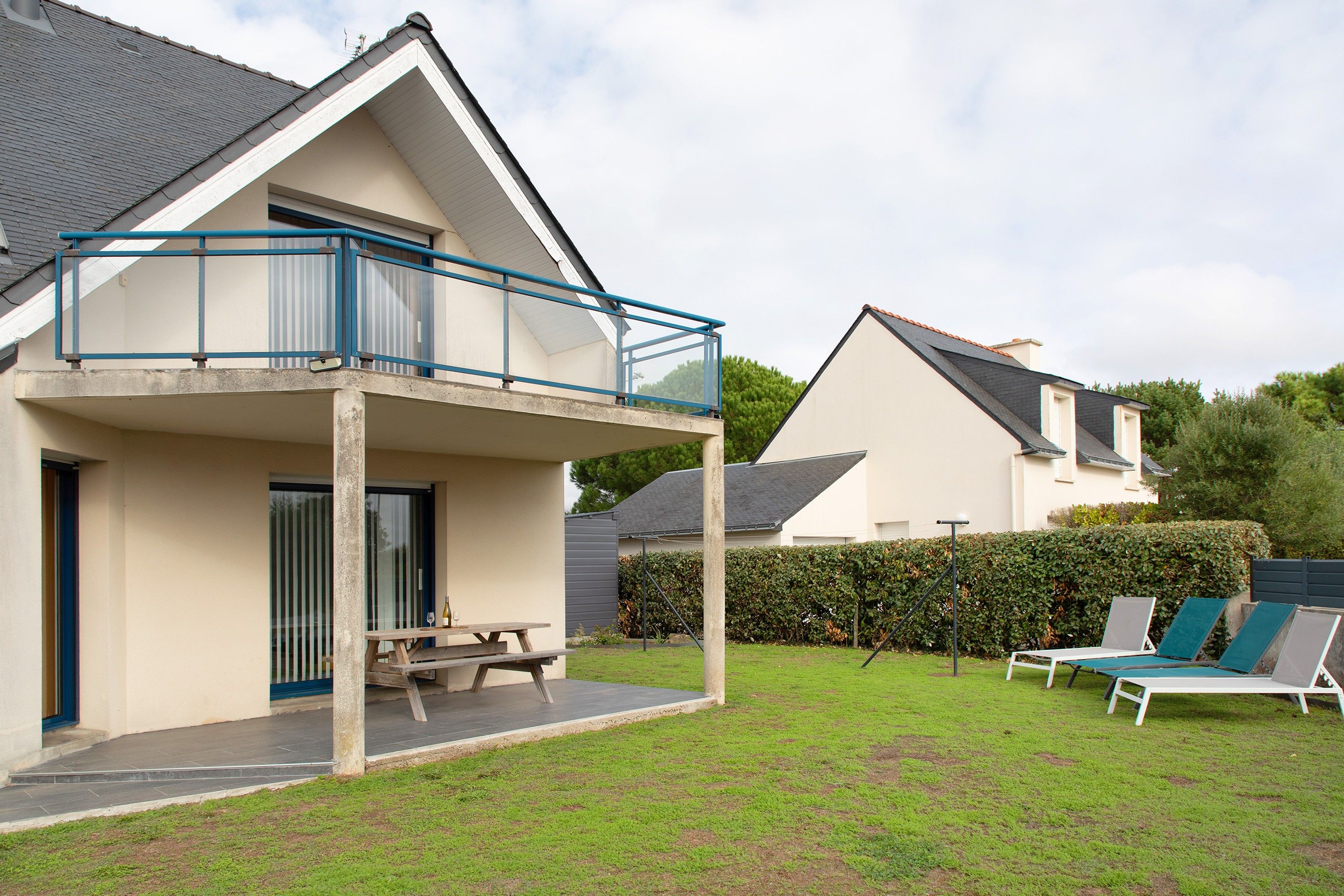 A two-story house with a balcony, a patio with a table and chairs, and a lawn with lounge chairs.
