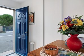 Una habitación con una puerta azul, un cuadro en la pared, una mesa de madera con un florero rojo y una cesta, y una ventana con vistas al exterior.