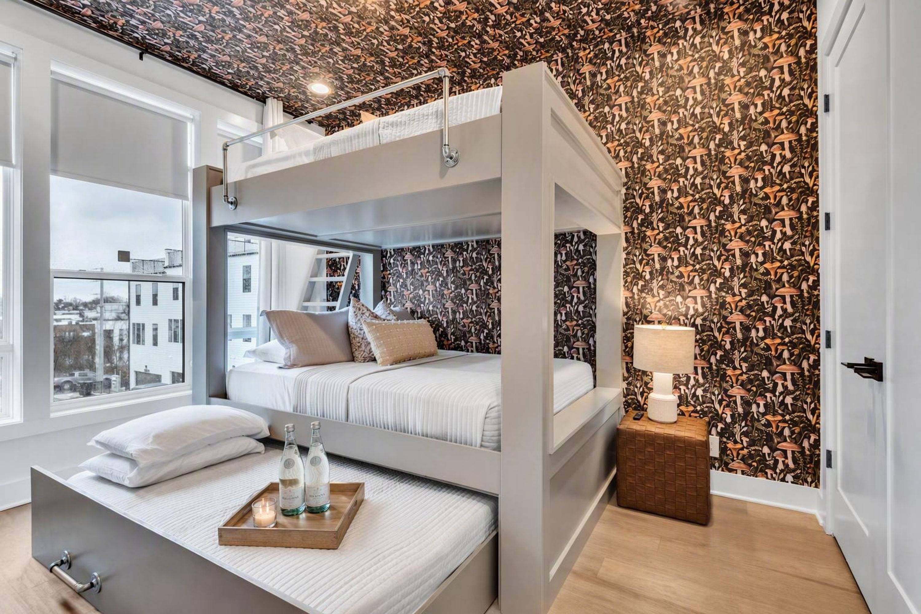 A modern bedroom with a bunk bed, a bedside table with a lamp, and a view of the city through large windows.