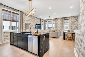 A modern kitchen with a central island, a dining area with a wooden table and chairs, and a living area with a sofa and a flat-screen TV. - 2 Homes - Rooftop Decks - Fire Pit - Sleeps 24 (Nashville)