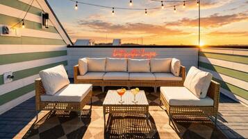 A rooftop terrace with a sofa set, two armchairs, a coffee table, and string lights against a sunset backdrop.