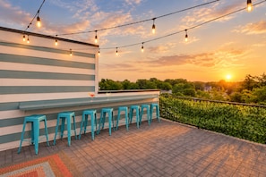 A rooftop bar with a row of blue stools, a bar counter, and string lights against a sunset backdrop. - 2 Homes - Rooftop Decks - Fire Pit - Sleeps 24 (Nashville)