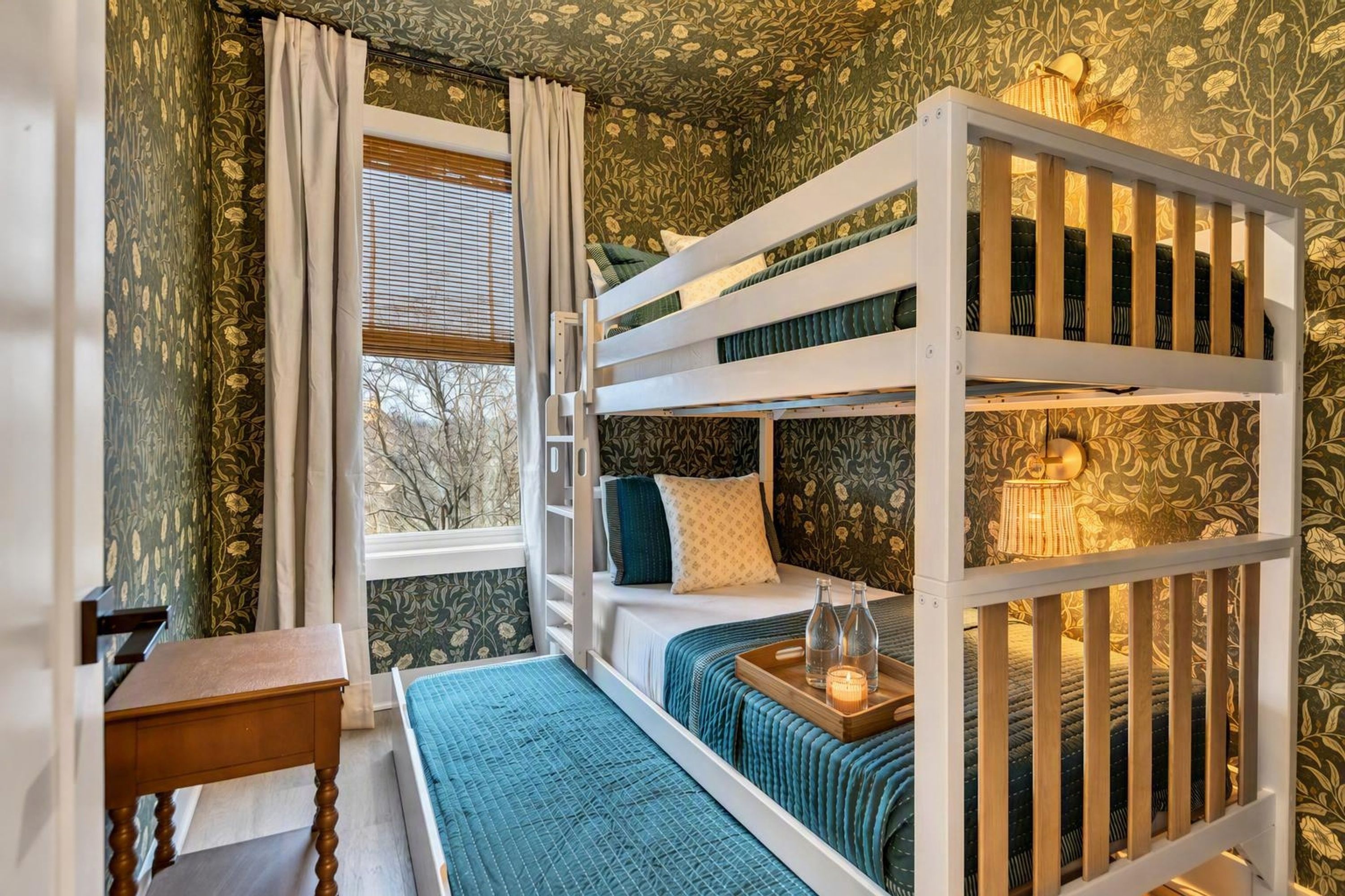 A bunk bed room with a window, a bedside table, and a lamp.