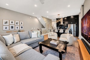 A modern living room with a grey sofa, a coffee table, and a TV screen displaying a concert. - 2 Homes - Rooftop Decks - Fire Pit - Sleeps 24 (Nashville)