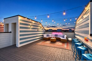 Una terraza en la azotea con un sofá curvo, taburetes azules de bar y luces colgantes.