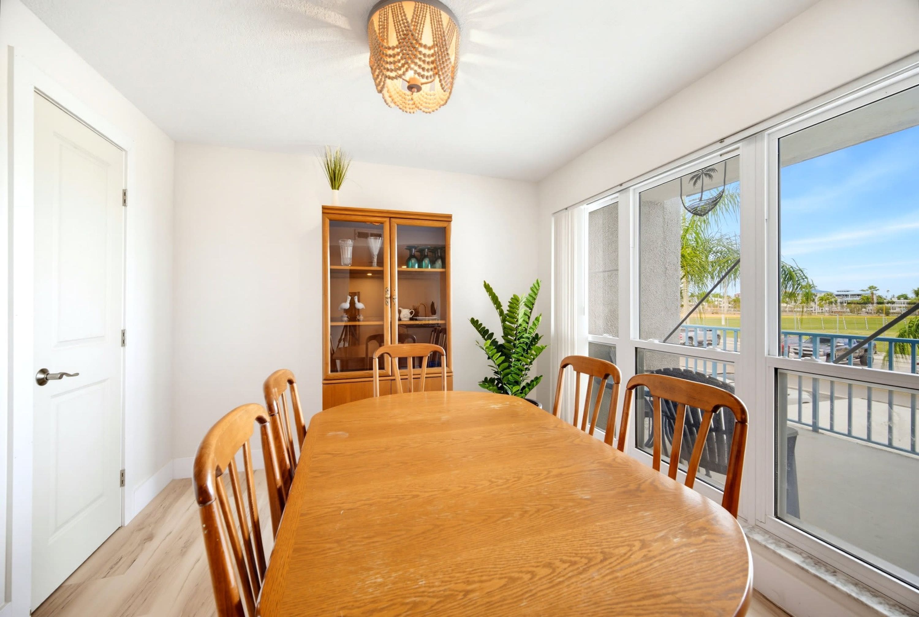 A dining room with a wooden table and chairs, a glass cabinet, and a view through large windows.