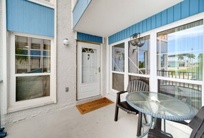 A covered patio area with a glass table and chairs, a door with a 'F' marking, and a welcome mat. - Walk to Beach Heated Pool - Madeira Beach 2BR (Madeira Beach)