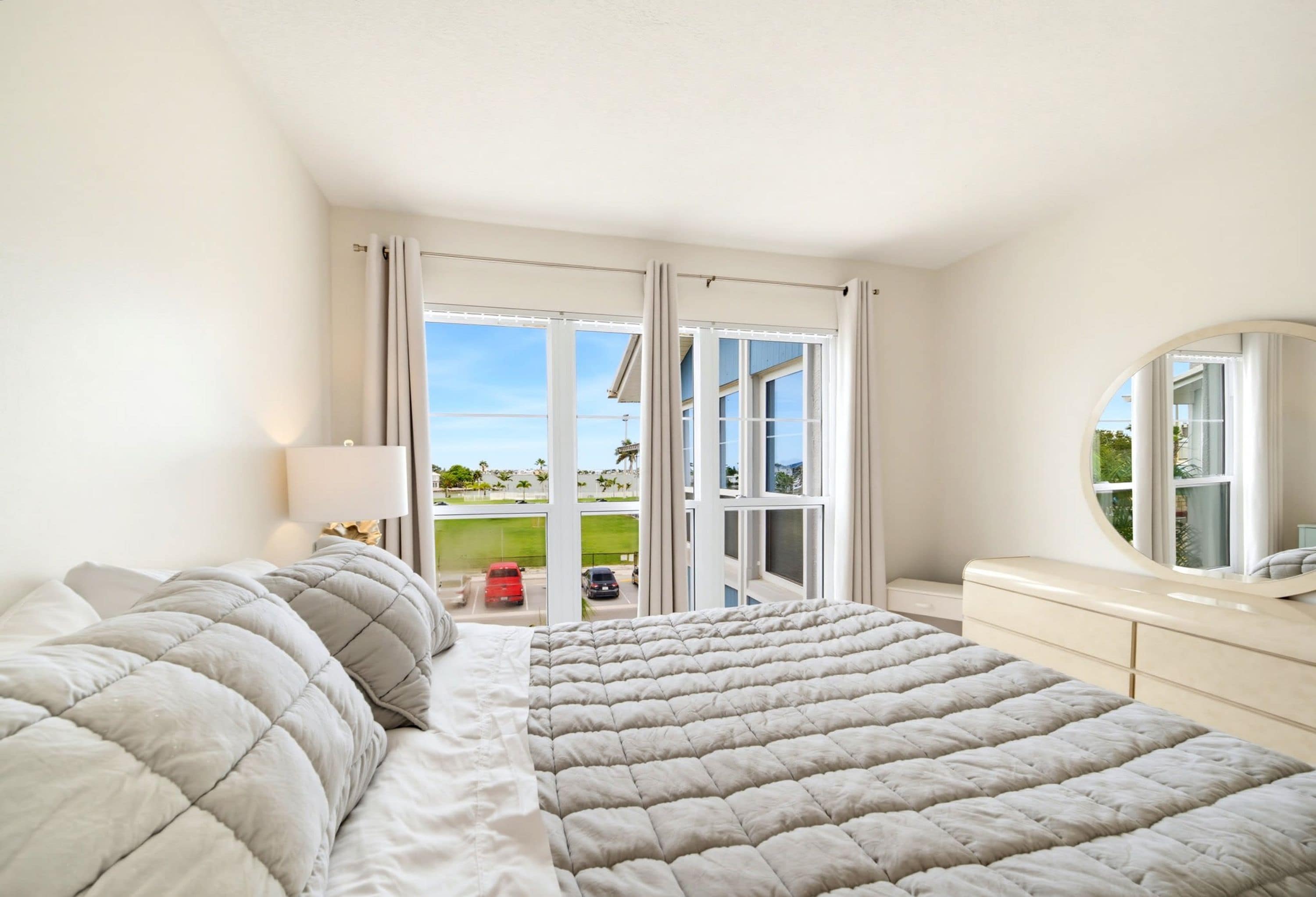 A bedroom with a large bed, a bedside table, a mirror, and a view of a parking area through a sliding door.