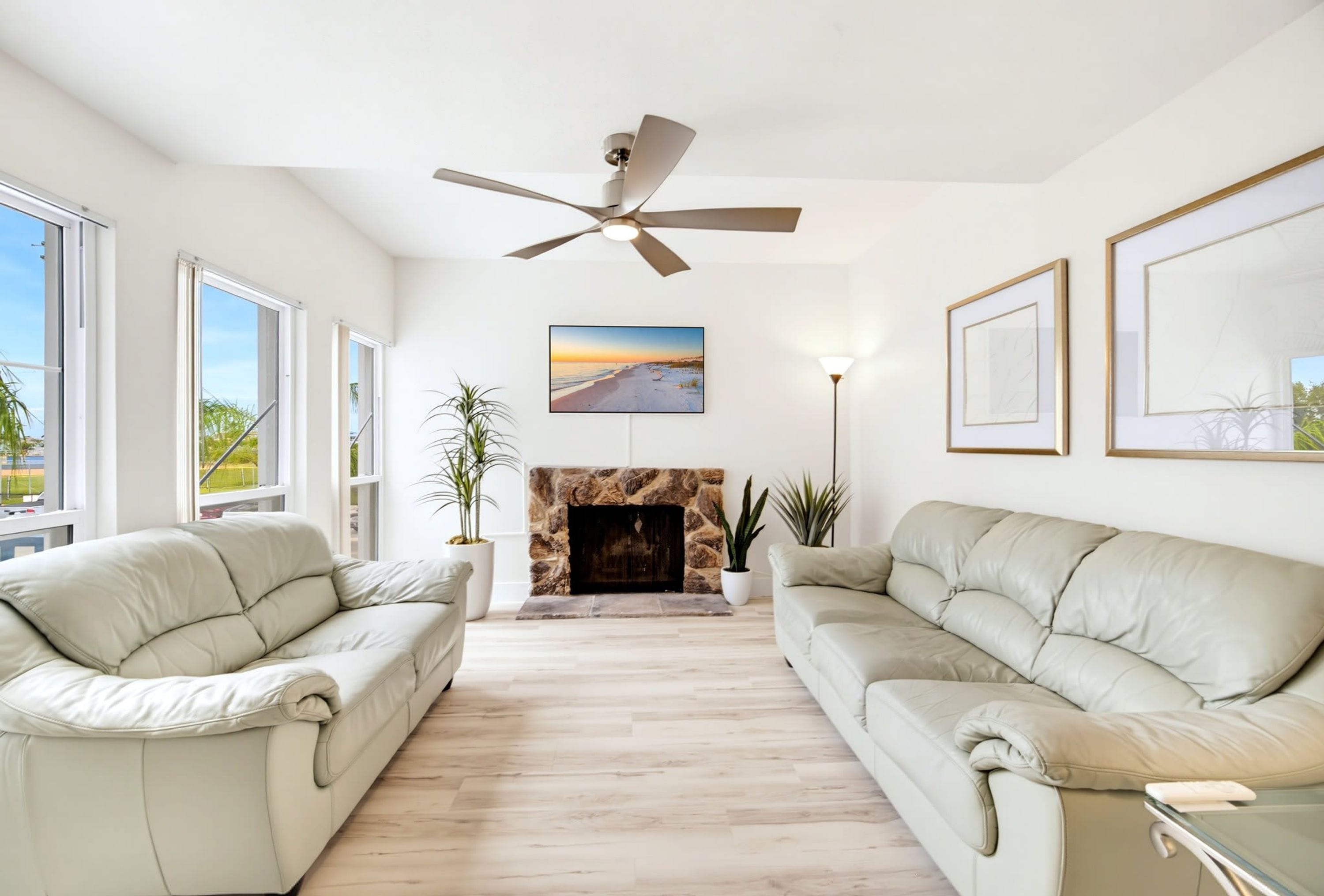 A spacious living room with two beige sofas, a stone fireplace, a ceiling fan, and large windows.