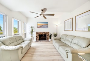 A spacious living room with two beige sofas, a stone fireplace, a ceiling fan, and large windows. - Walk to Beach Heated Pool - Madeira Beach 2BR (Madeira Beach)