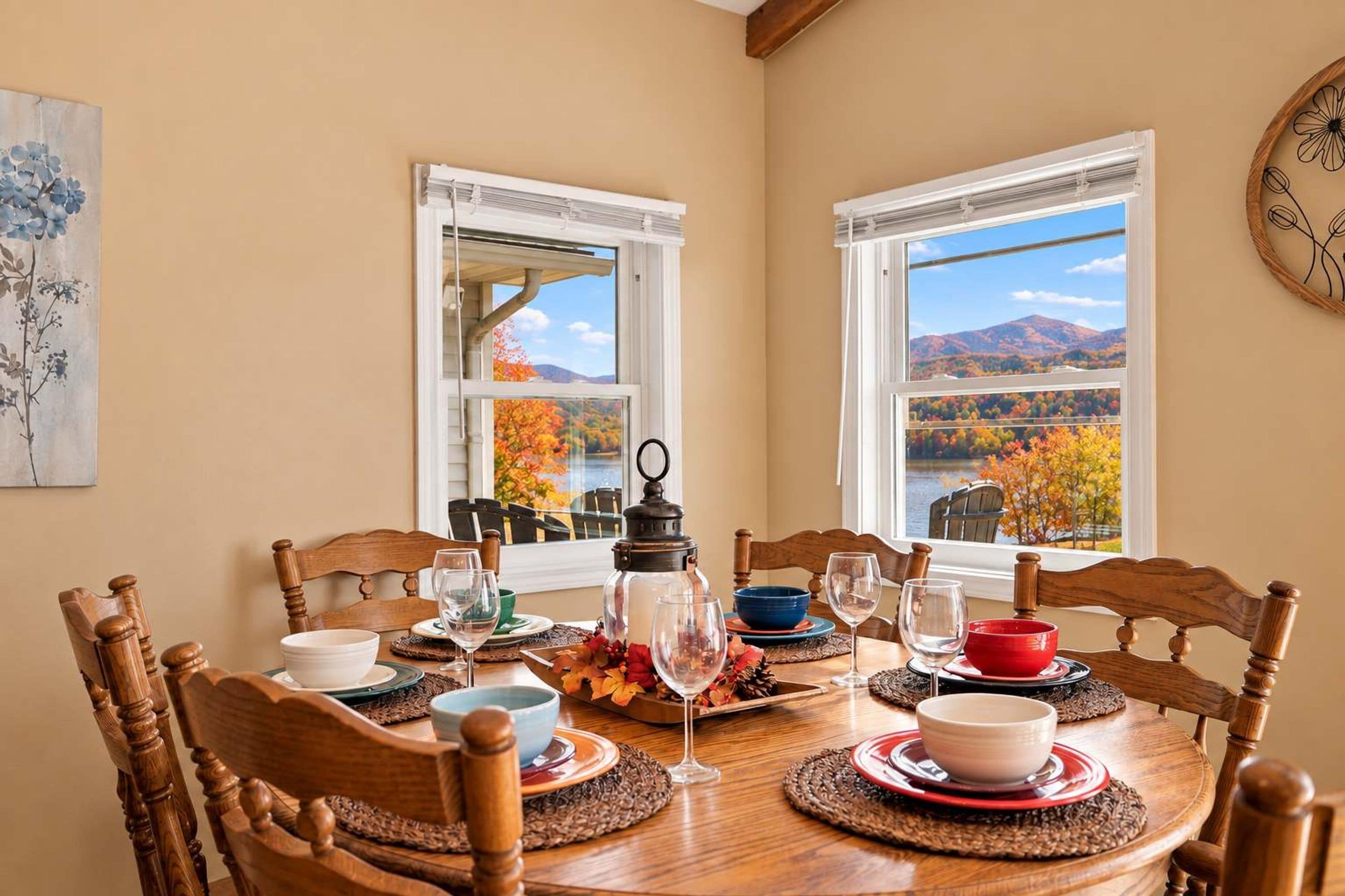 A dining room with a round table set for four, wooden chairs, and a view of autumn foliage through large windows.