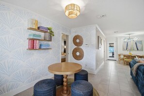 A modern interior with a round wooden table, blue woven ottomans, and a bookshelf with books and decorative items. - FWB Paradise Sports Court Hot Tub Putt Putt (Fort Walton Beach)