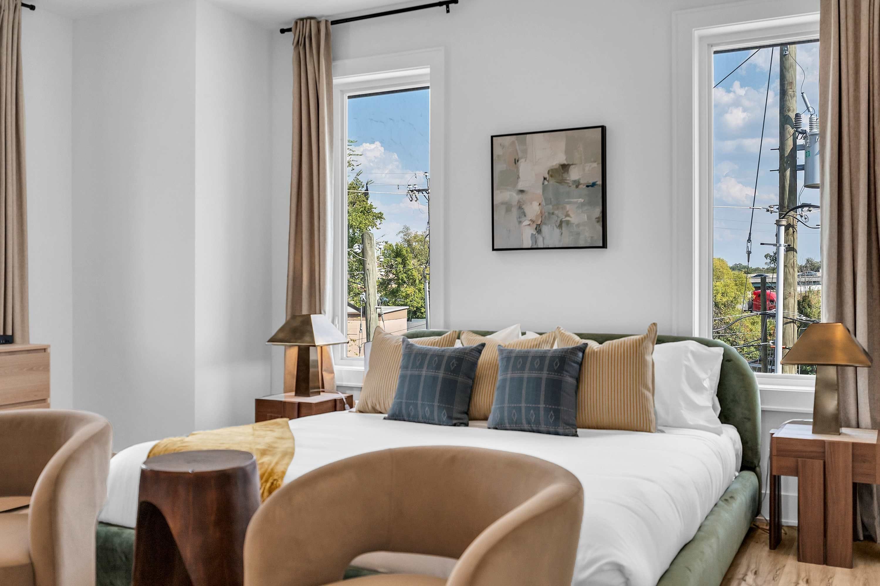 A modern bedroom with a large bed, multiple pillows, a side table, a chair, and a window with a view of the outside.