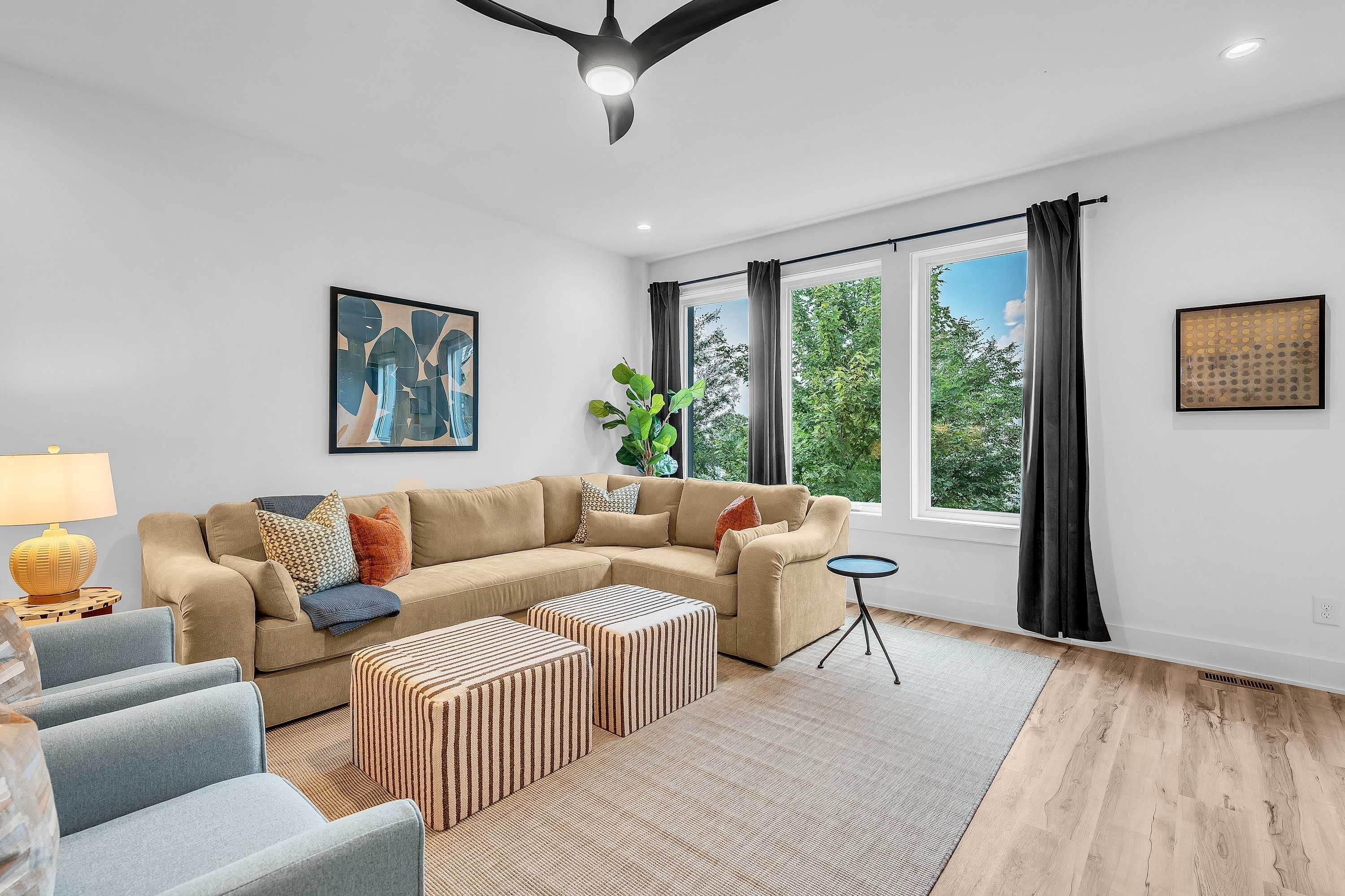 A modern living room with a sectional sofa, a coffee table, and a side table with a lamp. There are two windows with curtains, a ceiling fan, and a framed abstract painting on the wall.