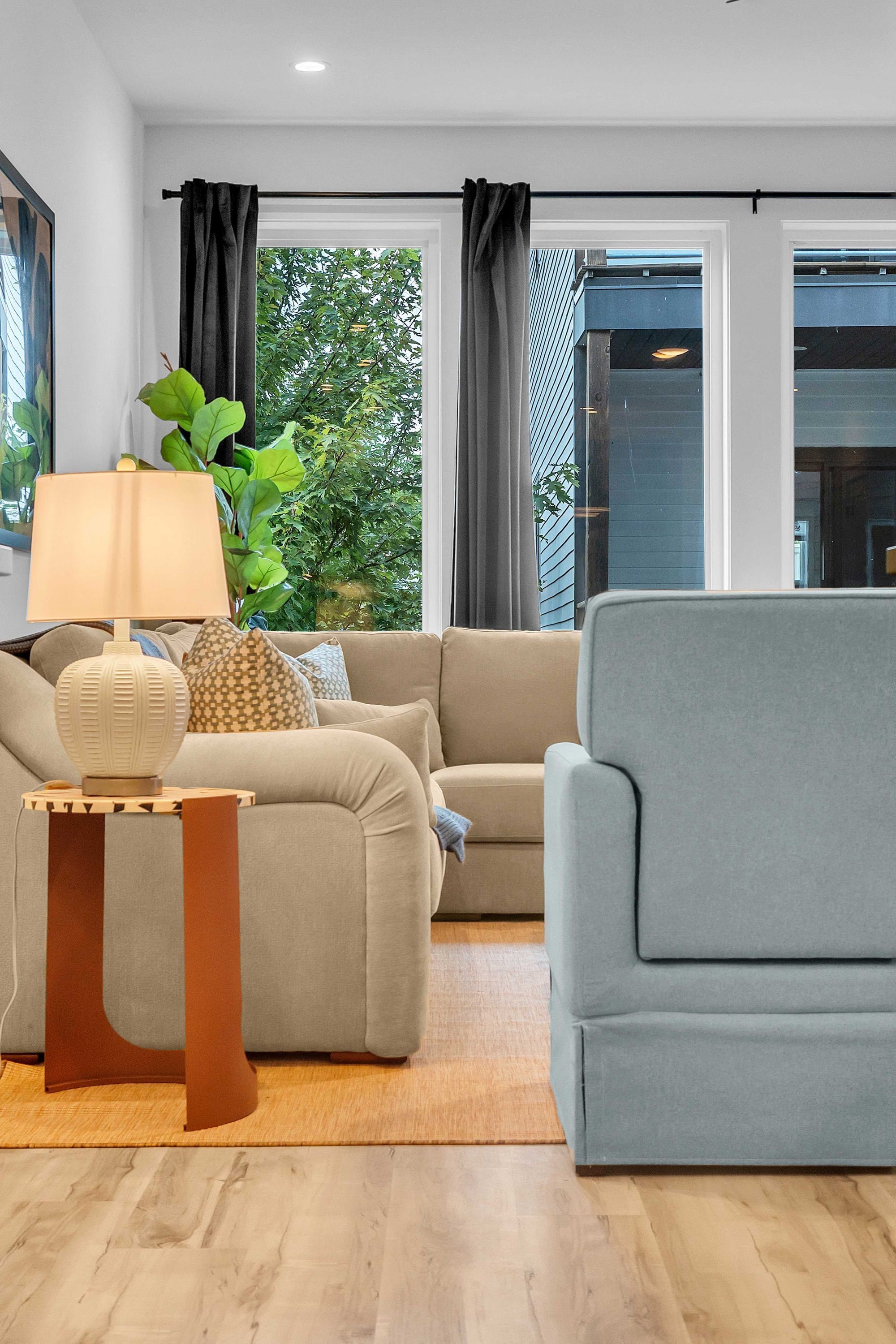 A modern living room with a sofa, a side table, a lamp, and a view of greenery outside.