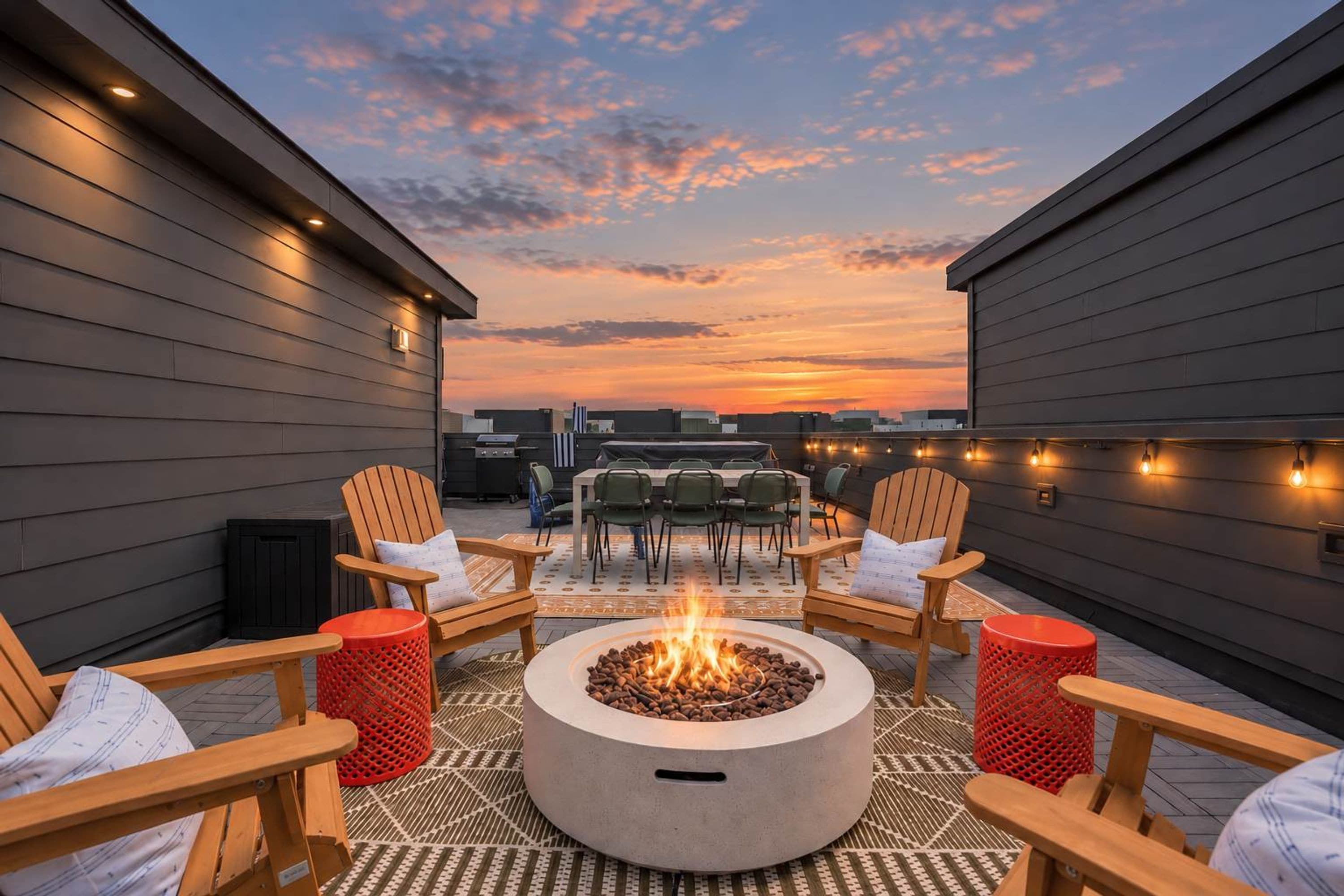 A rooftop patio with a central fire pit, surrounded by wooden chairs and tables, under a sunset sky.